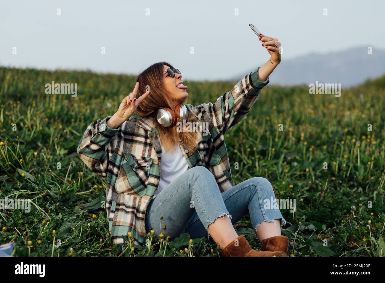Junge Frau in Sonnenbrillen mit Kopfhörern, die Friedensgesten zeigen und Die Zunge herausstreckt, während Sie Selbstporträts auf dem Handy und machen Sitzen auf der Wiese Stockfoto