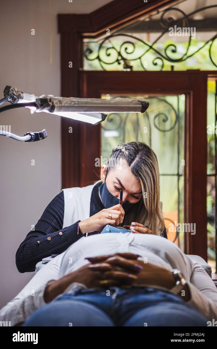 Kosmetologe mit Pinzette Anwendung gefälschte Wimpern zur Verlängerung auf das Auge Der ethnischen Klientin mit Gesichtsschutzmaske im Salon während Coronavirus-Pandem Stockfoto