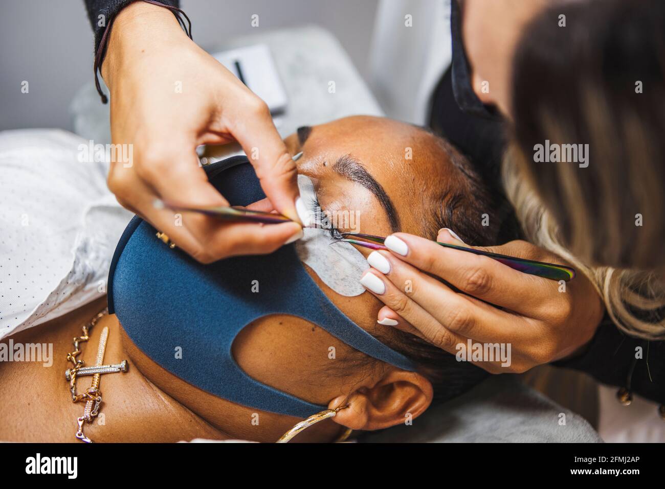 Hoher Winkel der Ernte unkenntlich Kosmetologe mit Pinzette Anwendung gefälschte Wimpern zur Erweiterung auf das Auge des ethnischen Kunden mit Gesicht Schutzmaske Stockfoto