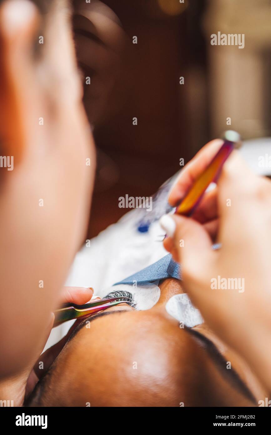 Crop unkenntlich Kosmetologe mit Pinzette Anwendung gefälschte Wimpern zur Verlängerung Auf das Auge eines ethnischen Kunden mit Gesichtsschutzmaske in Salon durin Stockfoto