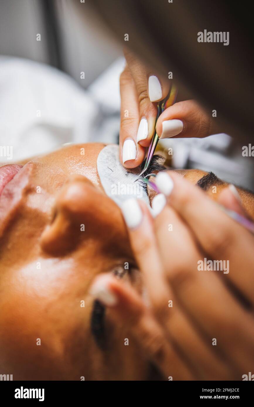 Hoher Winkel der Ernte unkenntlich Kosmetologe mit Pinzette Anwendung gefälschte Wimpern zur Erweiterung auf das Auge des ethnischen Kunden im Salon Stockfoto
