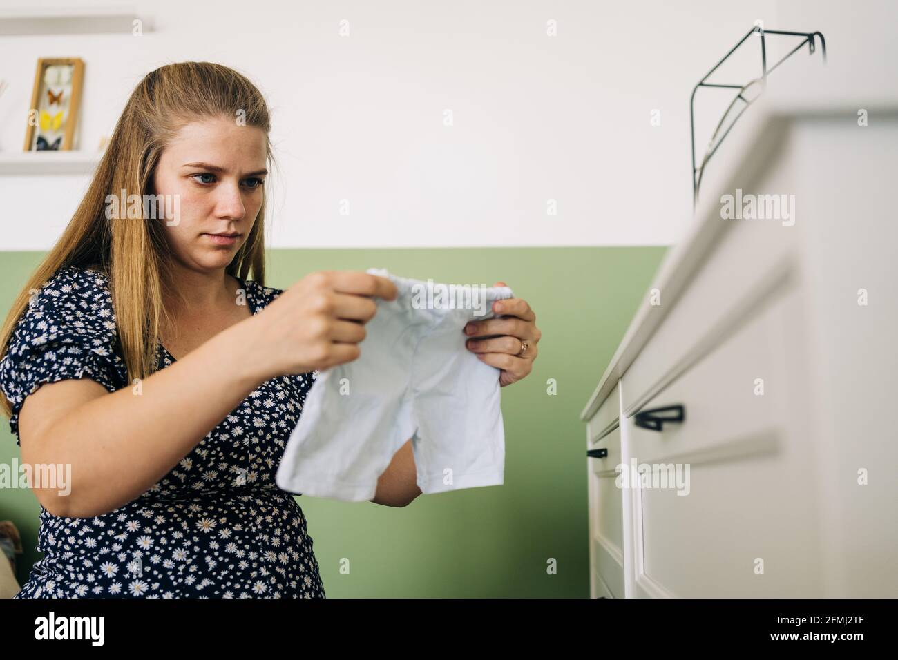 Werdende Frau in Zierkleid sitzt auf Yoga-Ball mit Kinderkleidung gegen Kommode im Zimmer Stockfoto