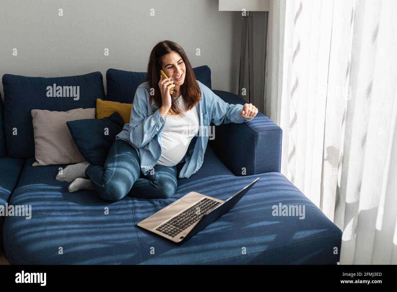 Schwangerin sitzt auf dem Sofa mit Laptop im Wohnzimmer Raum und sprechen auf dem Handy, während Remote-Projekt zu diskutieren Und arbeiten von ho Stockfoto