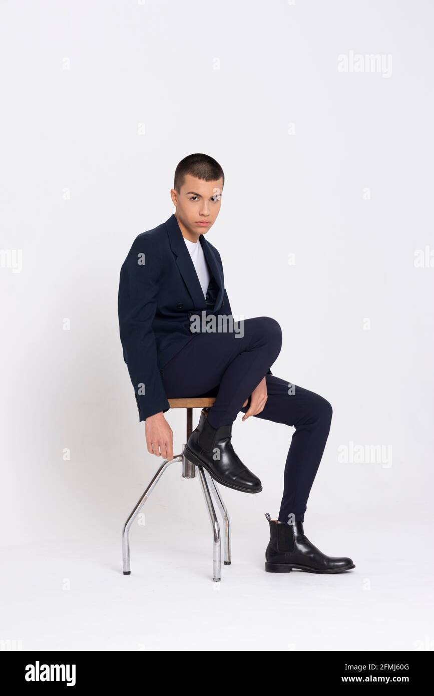 Seitenansicht des entschlossenen Mannes mit elegantem Anzug im Sitzen Hocker auf weißem Hintergrund im Studio und Blick auf die Kamera Stockfoto