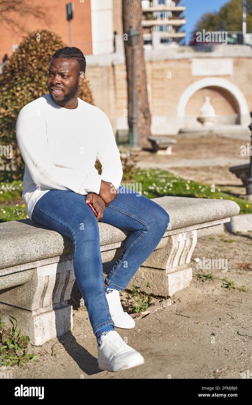 Glücklicher afroamerikanischer Mann, der in einem Park sitzt und Jeans,  weißen Pullover und weißen Turnschuhen trägt. Lifestyle-Konzept. Hochwertige  Fotos Stockfotografie - Alamy