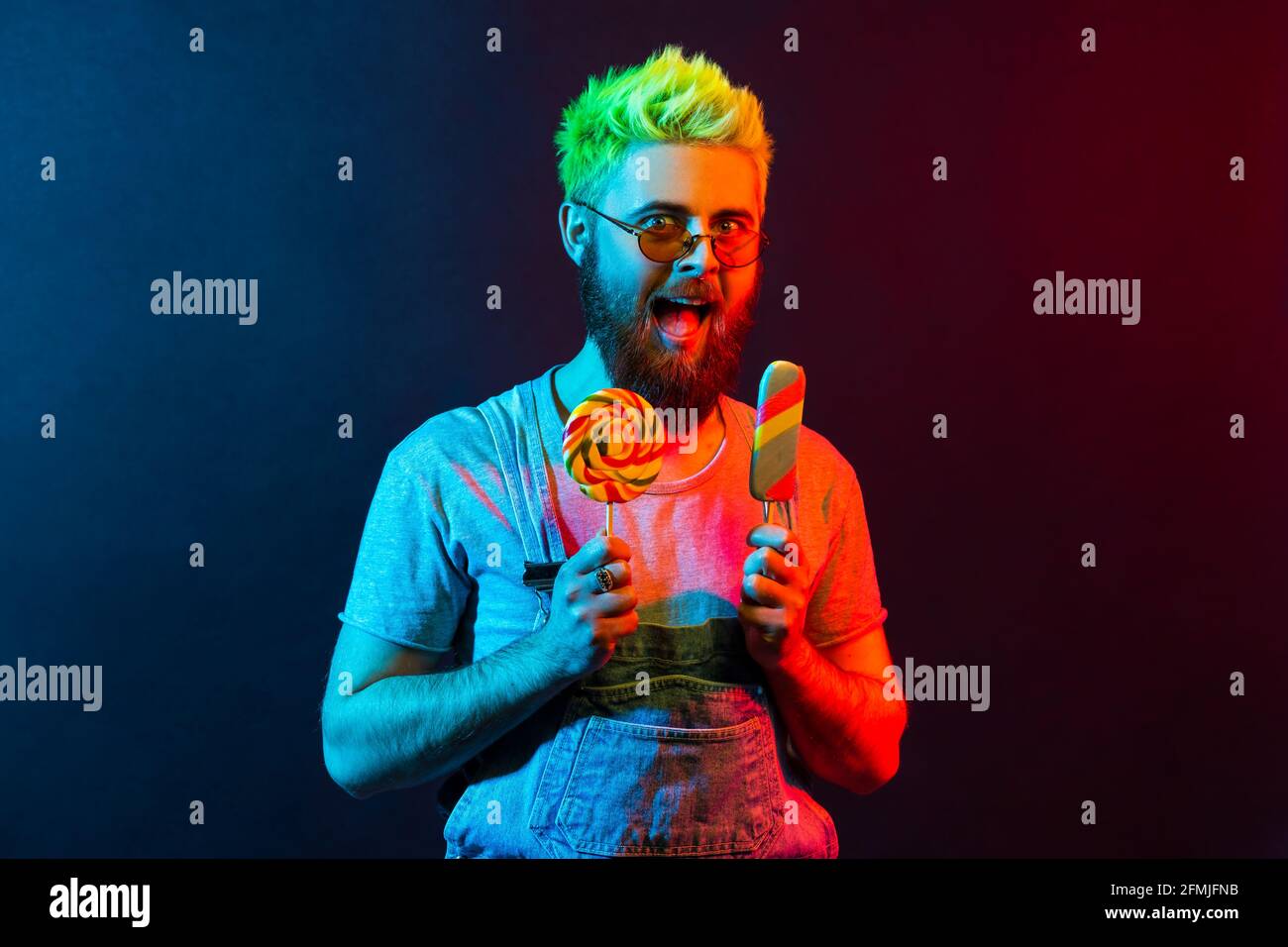 Extrem fröhlicher Hipster-Typ in Denim insgesamt, der Lollipop und Eis in den Händen hält, vor Aufregung schreit, verrückt nach süßem zuckerhaltigem Konfekt Stockfoto