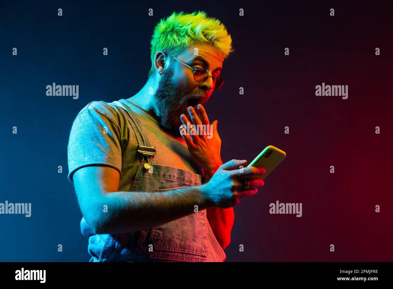 Junger Erwachsener attraktiver Hipster Kerl mit Bart mit Handy in der Hand sieht etwas Erstaunliches auf dem Display, bedeckt weit geöffneten Mund mit Handfläche. C Stockfoto
