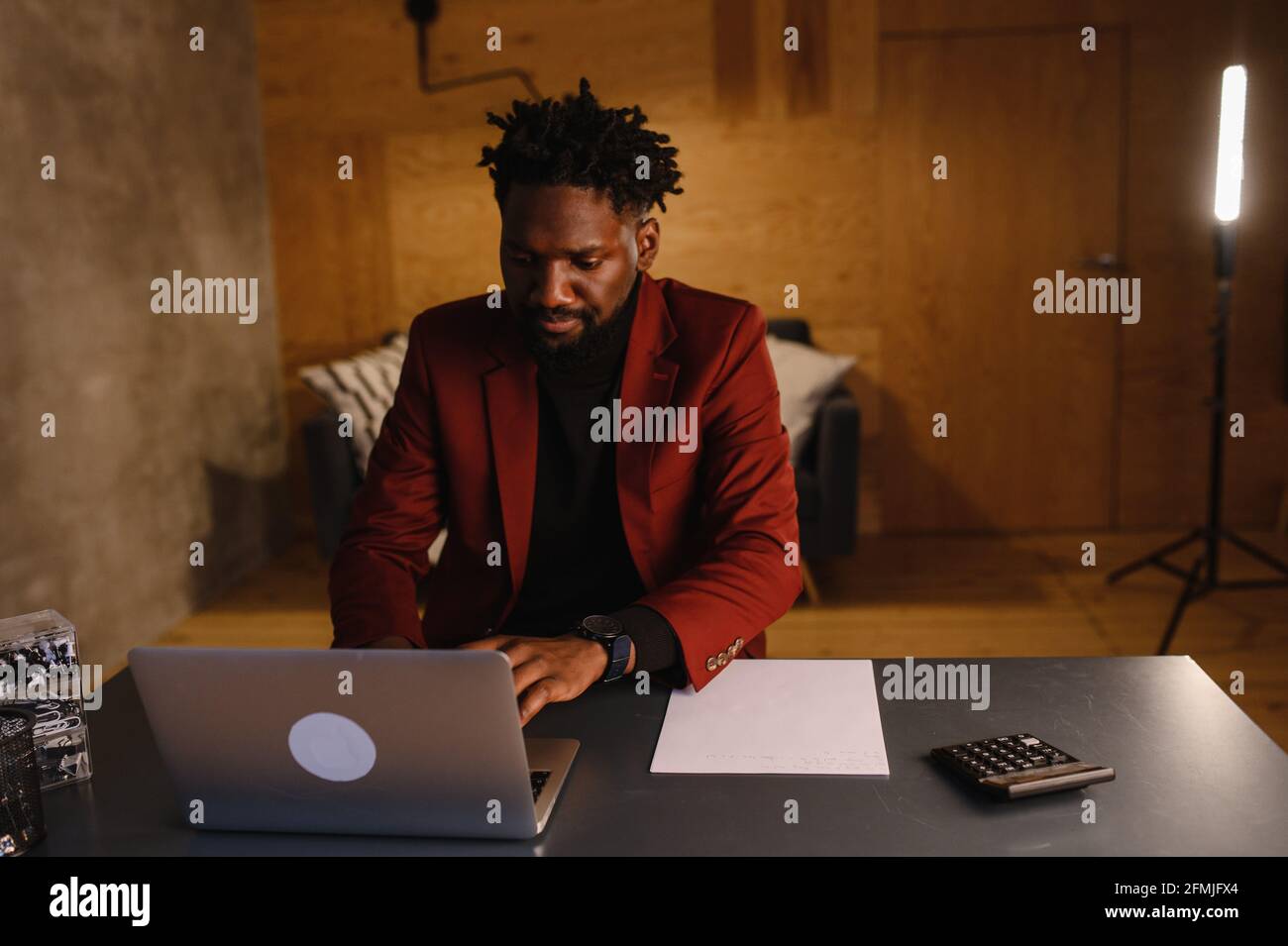 Schöner schwarzer afroamerikanischer Mann, der am Laptop-Computer arbeitet, während er hinter dem Schreibtisch im gemütlichen Wohnzimmer sitzt. Freiberuflicher Mitarbeiter, Der Von Zu Hause Aus Arbeitet Stockfoto