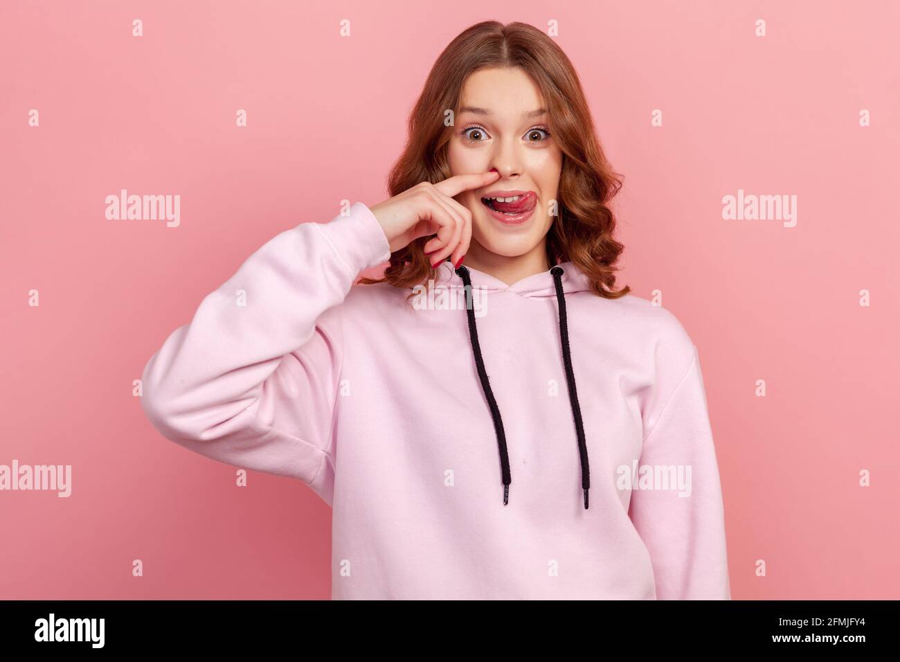 Porträt von lustigen Teenager lockig haarige Mädchen in Hoodie bohren Nase herumtollend, zeigt Zunge aus und Kommissionierung Nase mit komisch dummen Ausdruck. Zoll Stockfoto