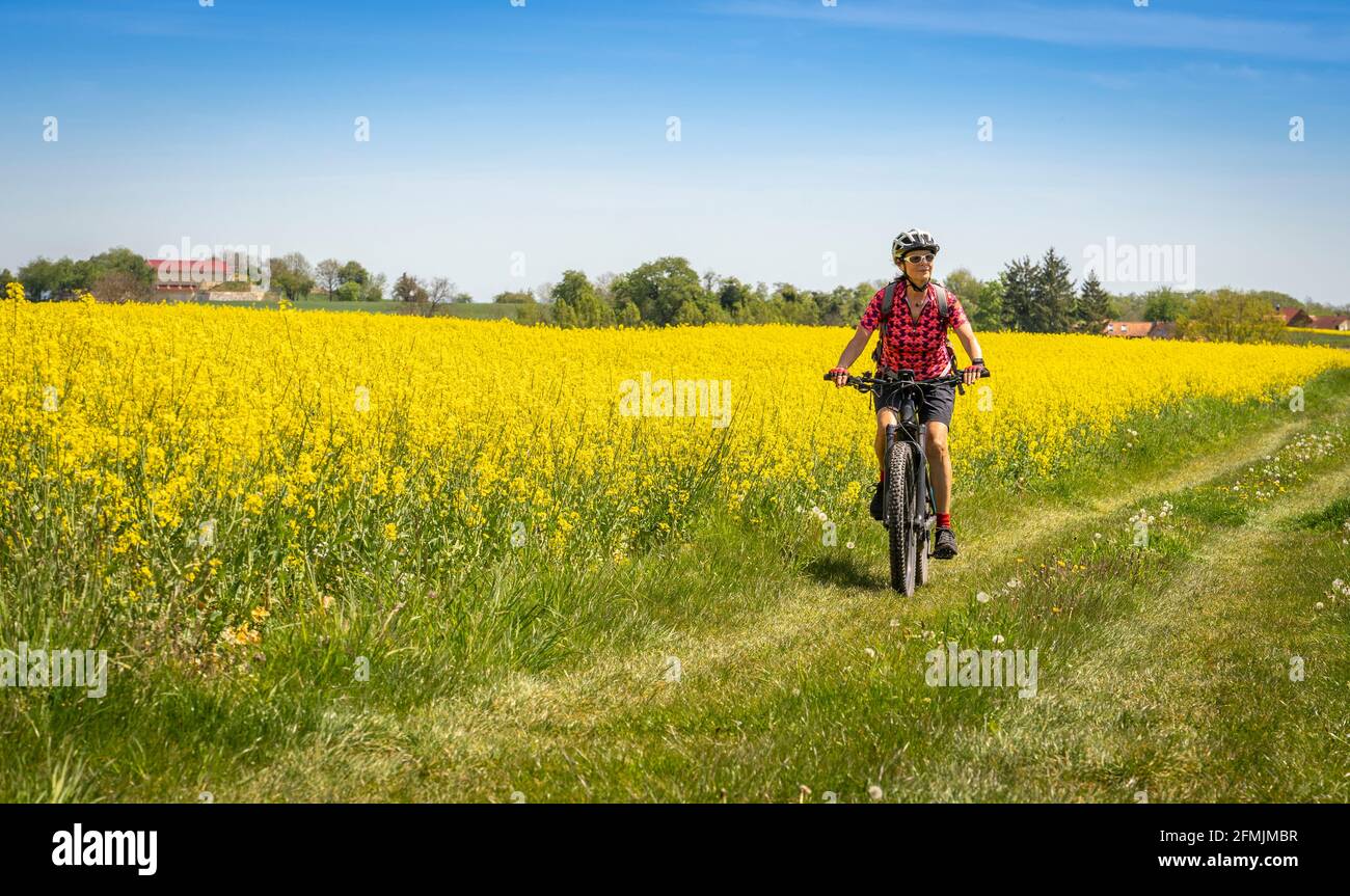 Nette und verbliebene junge ältere Frau auf dem Elektro-Mountainbike in einer Frühlingslandschaft zwischen Rapsfeldern im Kraichgau bei Zaberfeld, Bad Stockfoto