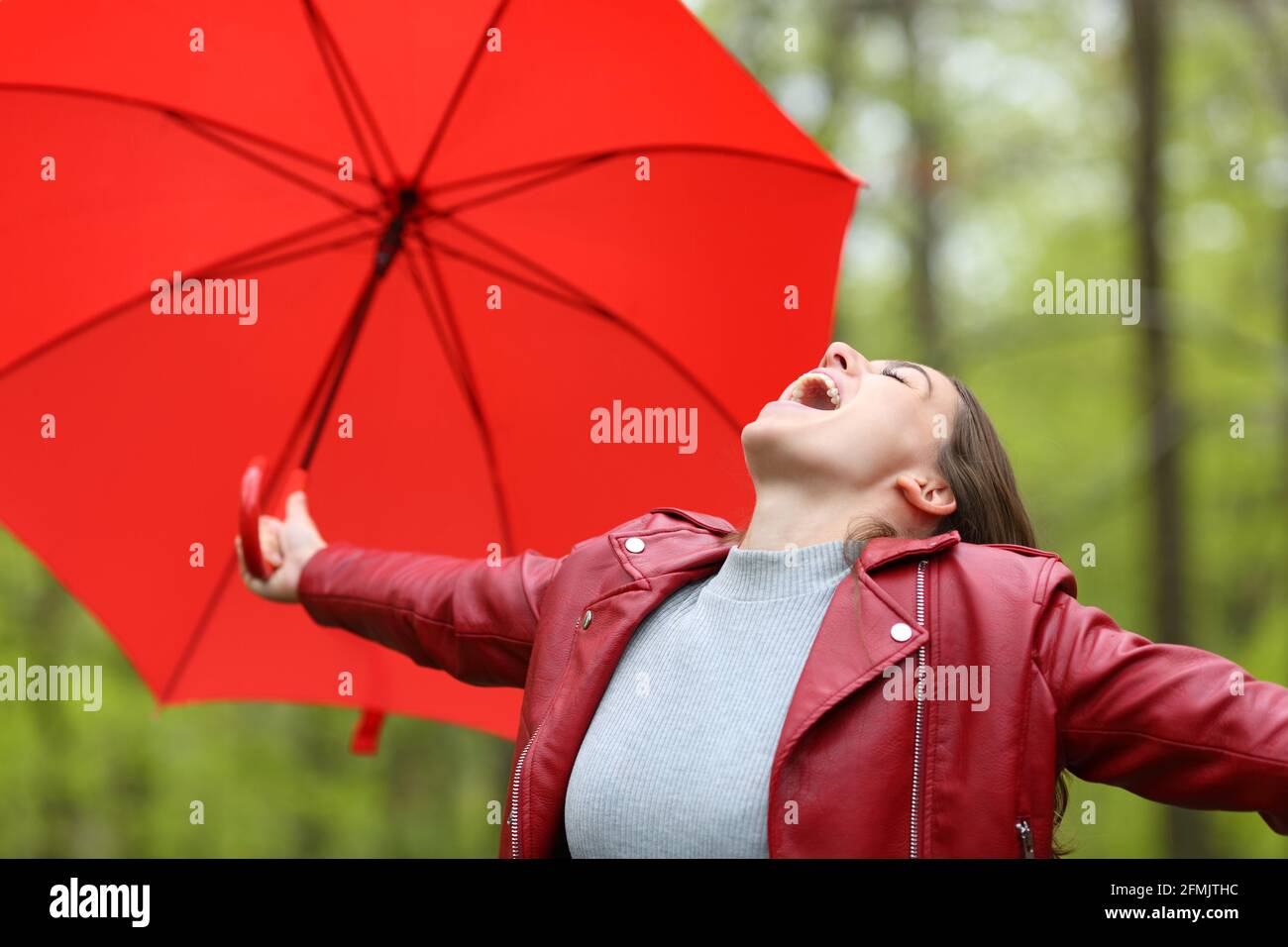Aufgeregte Frau in Rot feiert Glück unter dem Regen halten Regenschirm in einem Wald Stockfoto