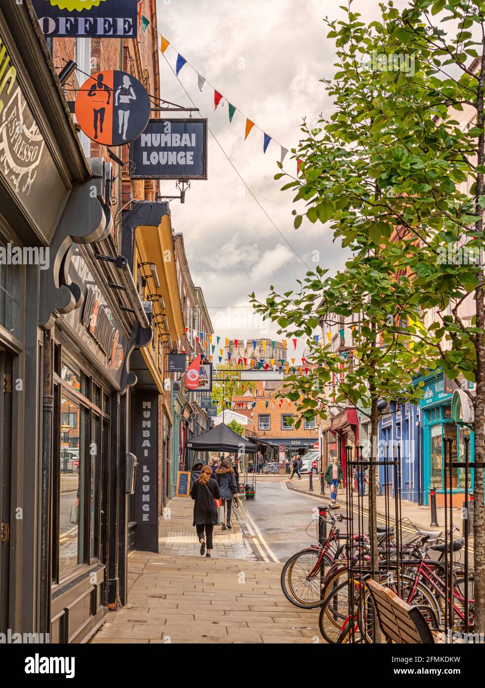 Eine Straße, die mit bunten Hahnenkästen befahren ist. Geschäfte und Restaurants befinden sich auf beiden Seiten. Fahrräder stehen im Vordergrund und eine Frau läuft auf dem Bürgersteig Stockfoto