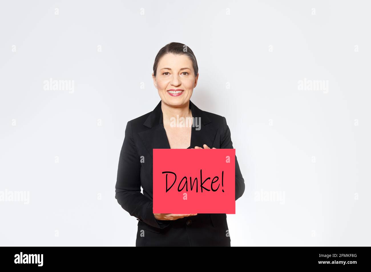 Lächelnde Frau mittleren Alters in schwarzem Blazer hält ein rotes Schild mit dem deutschen Text danke, copy space. Stockfoto