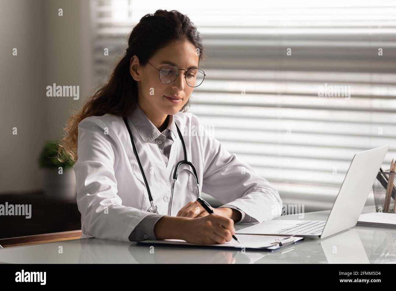 Konzentrierte hispanische Therapeutin sitzt am Schreibtisch und bereitet einen medizinischen Bericht vor Stockfoto