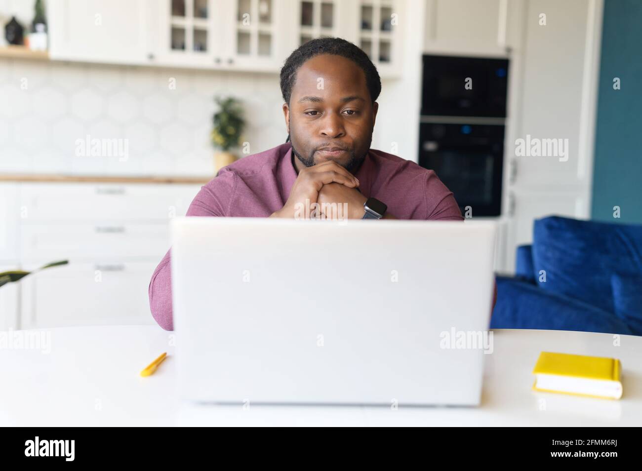 Fokussierter und besorgter schwarzer Mann in elegantem Casual-Shirt sieht auf den Laptop-Bildschirm, seriös afroamerikanischer Kerl sitzt am Schreibtisch, das Kinn auf den Händen ruht, fühlt Zweifel und löst schwierige Aufgaben Stockfoto