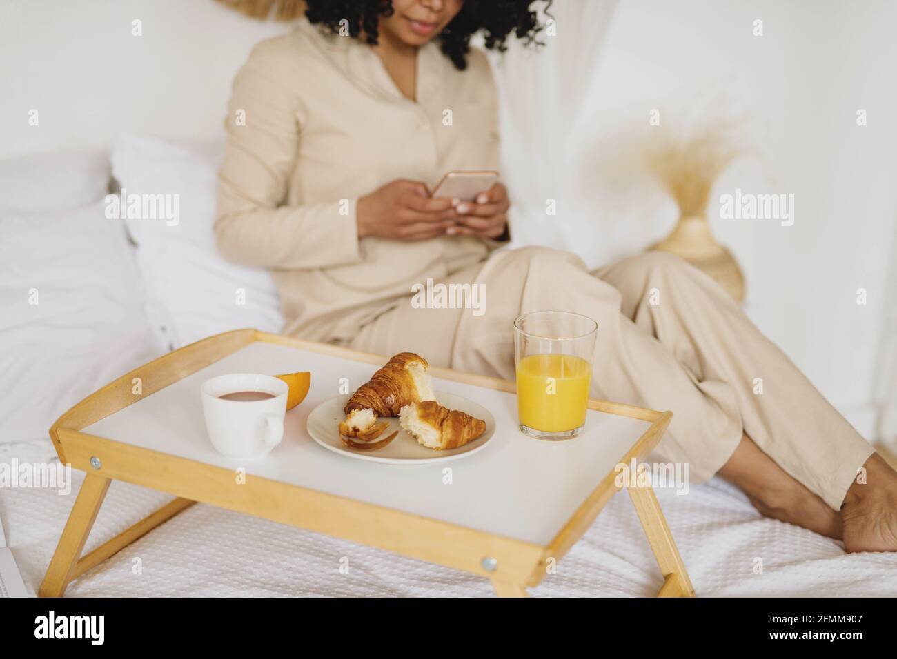 Junge afroamerikanische Frau mit Smartphone und Frühstück im Bett. Stockfoto
