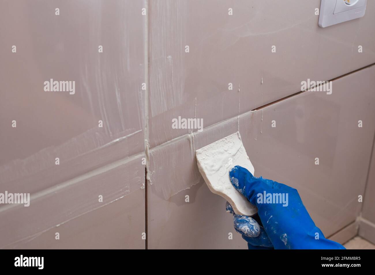 Reparatur und Wandverkleidung mit Fliesen. Der Meister reibt die Fliesenfugen mit einem weißen Kitt mit einem Gummispachtel. Stockfoto