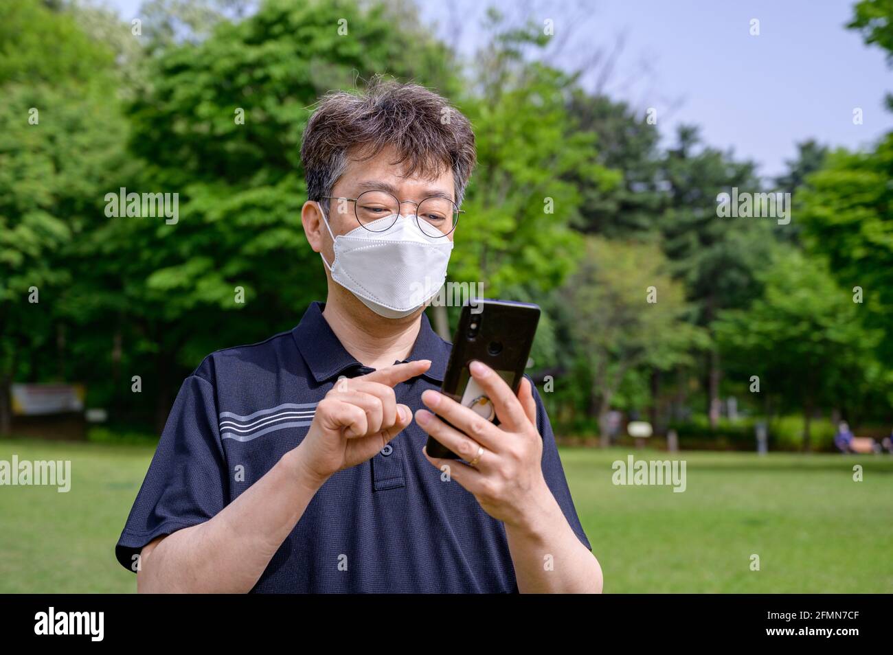 Ein asiatischer Mann mittleren Alters, der eine Gesichtsmaske trägt und ein Smartphone auf dem Rasen verwendet. Stockfoto