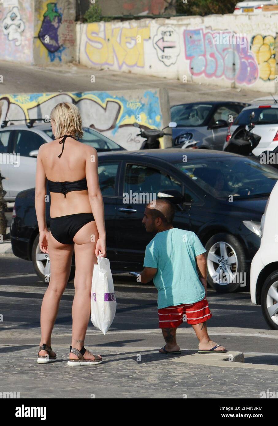 Ein Zwergmann, der neben einer blonden Frau im Bikini an der Uferpromenade in Tel-Aviv, Israel, steht. Stockfoto