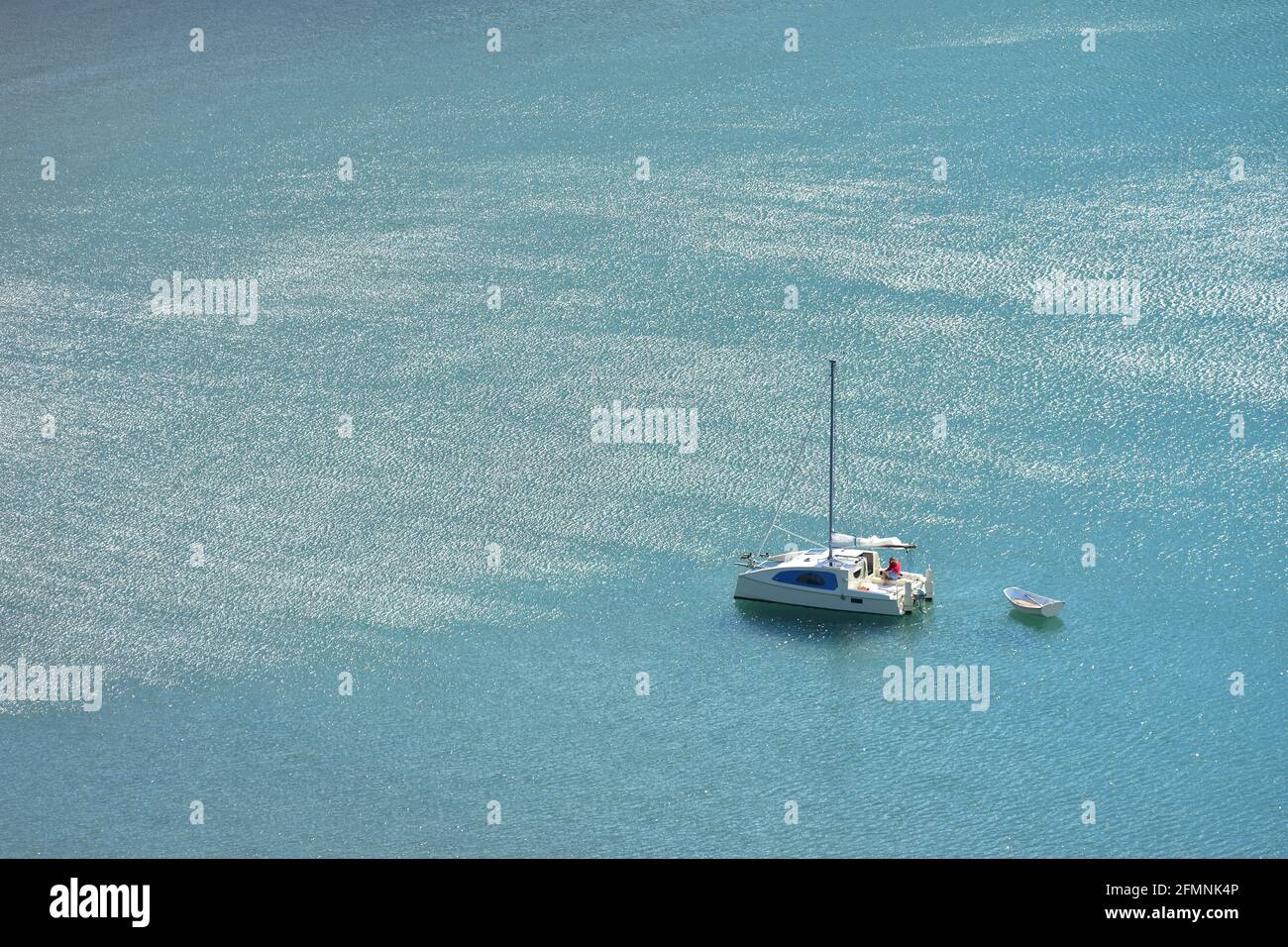 Kleiner Segel-Katamaran mit verstauten Segeln, der ein leichtes weißes Vintage-Schlauchboot auf dem ruhigen türkisfarbenen Meer vor Anker führt. Stockfoto