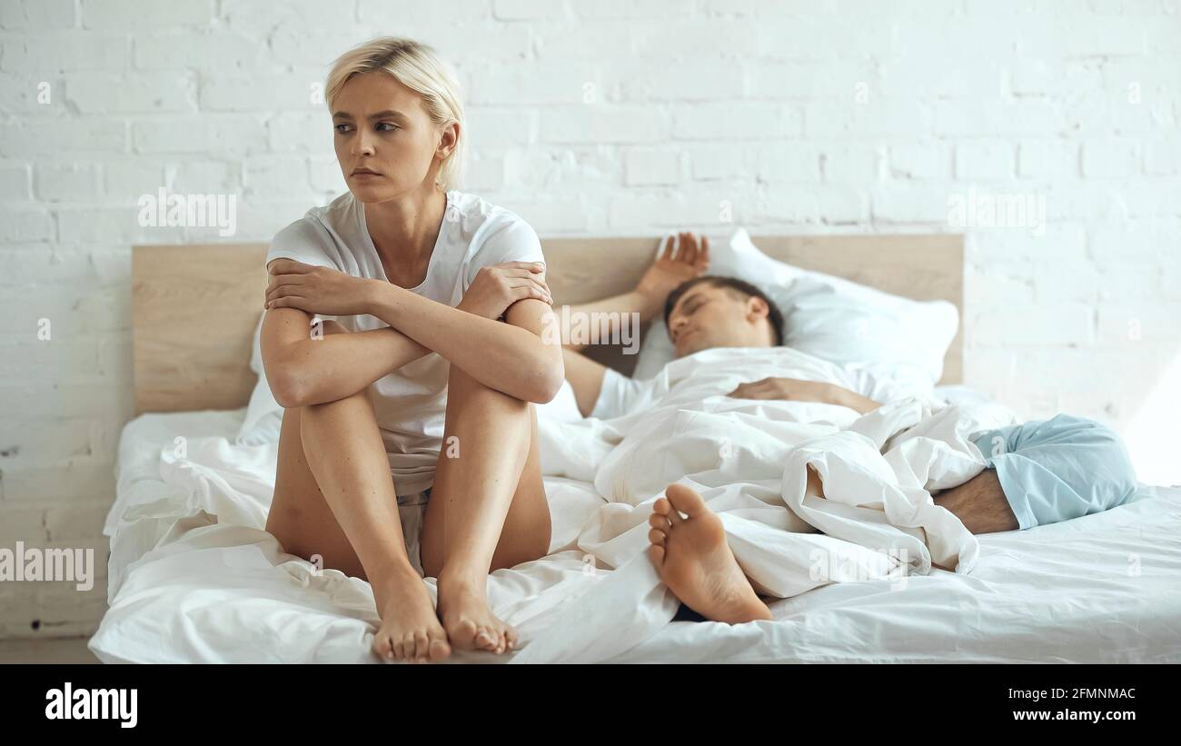 Traurige und barfuß sitzende Frau auf dem Bett, während der Freund schläft Im Schlafzimmer Stockfoto