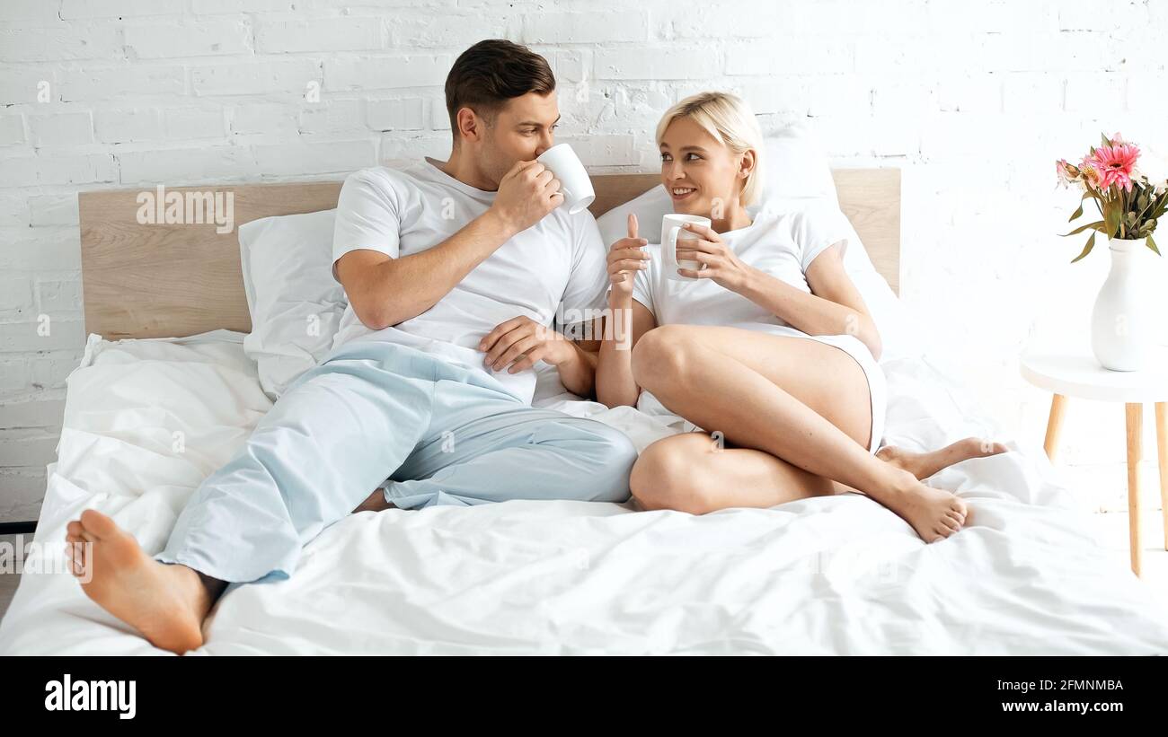 Lächelnde junge Frau, die den Freund beim Kaffeetrinken im Bett ansieht Stockfoto