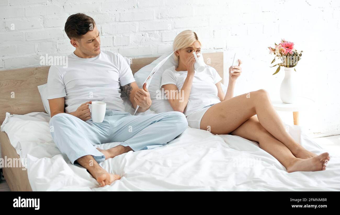 Tätowierte Mann Zeitung lesen in der Nähe Frau Kaffee trinken und verwenden Smartphone im Bett Stockfoto