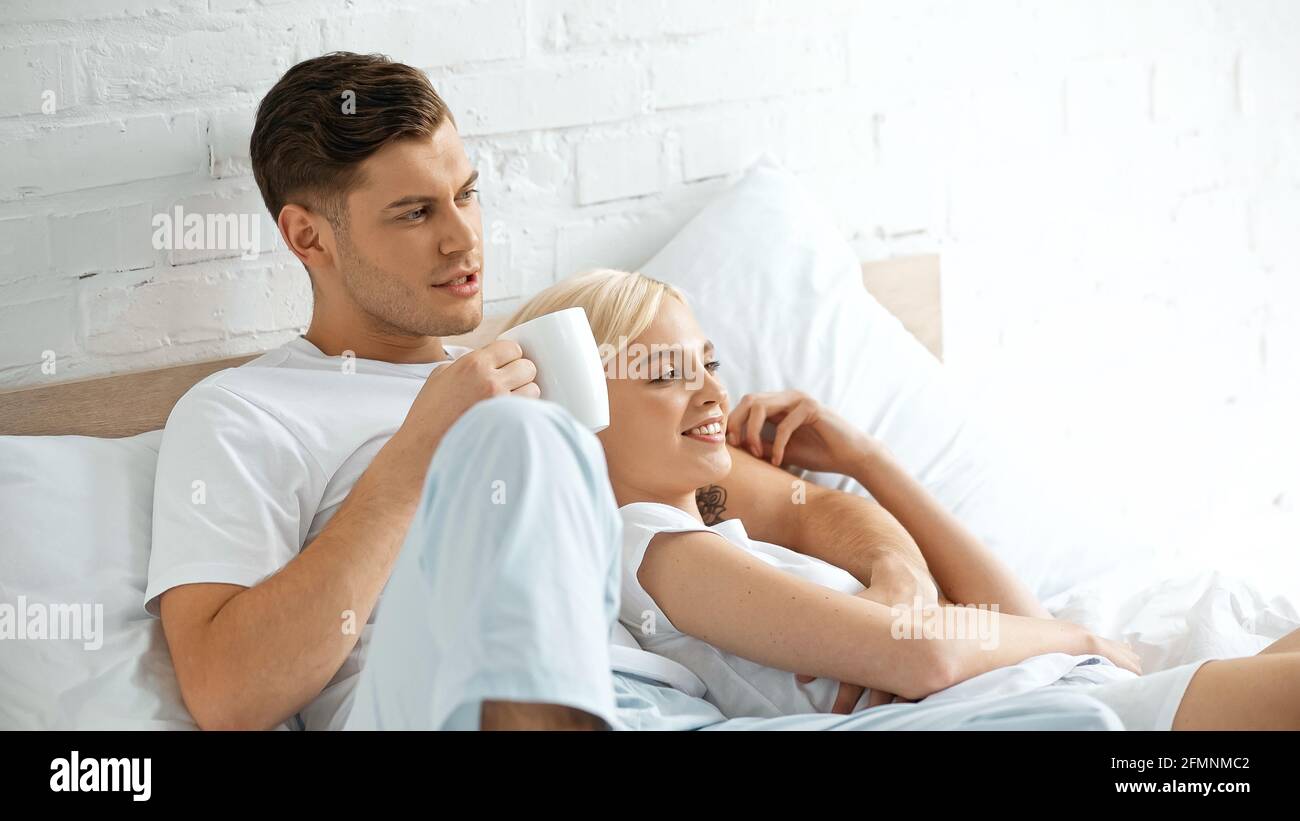 Tätowierte Mann hält Tasse Kaffee in der Nähe glückliche Frau liegend Im Bett Stockfoto