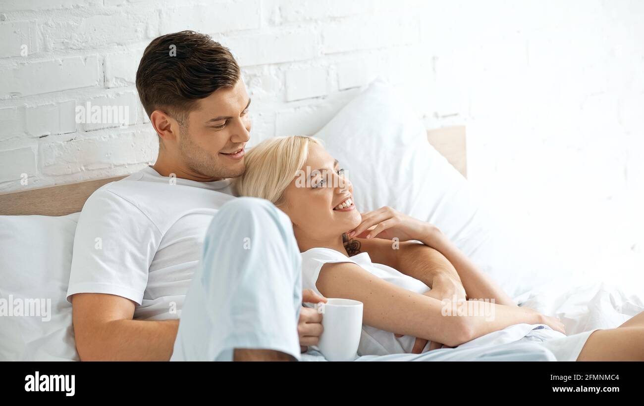 Tätowierte Mann hält Tasse Kaffee in der Nähe fröhliche Frau liegend Im Bett Stockfoto