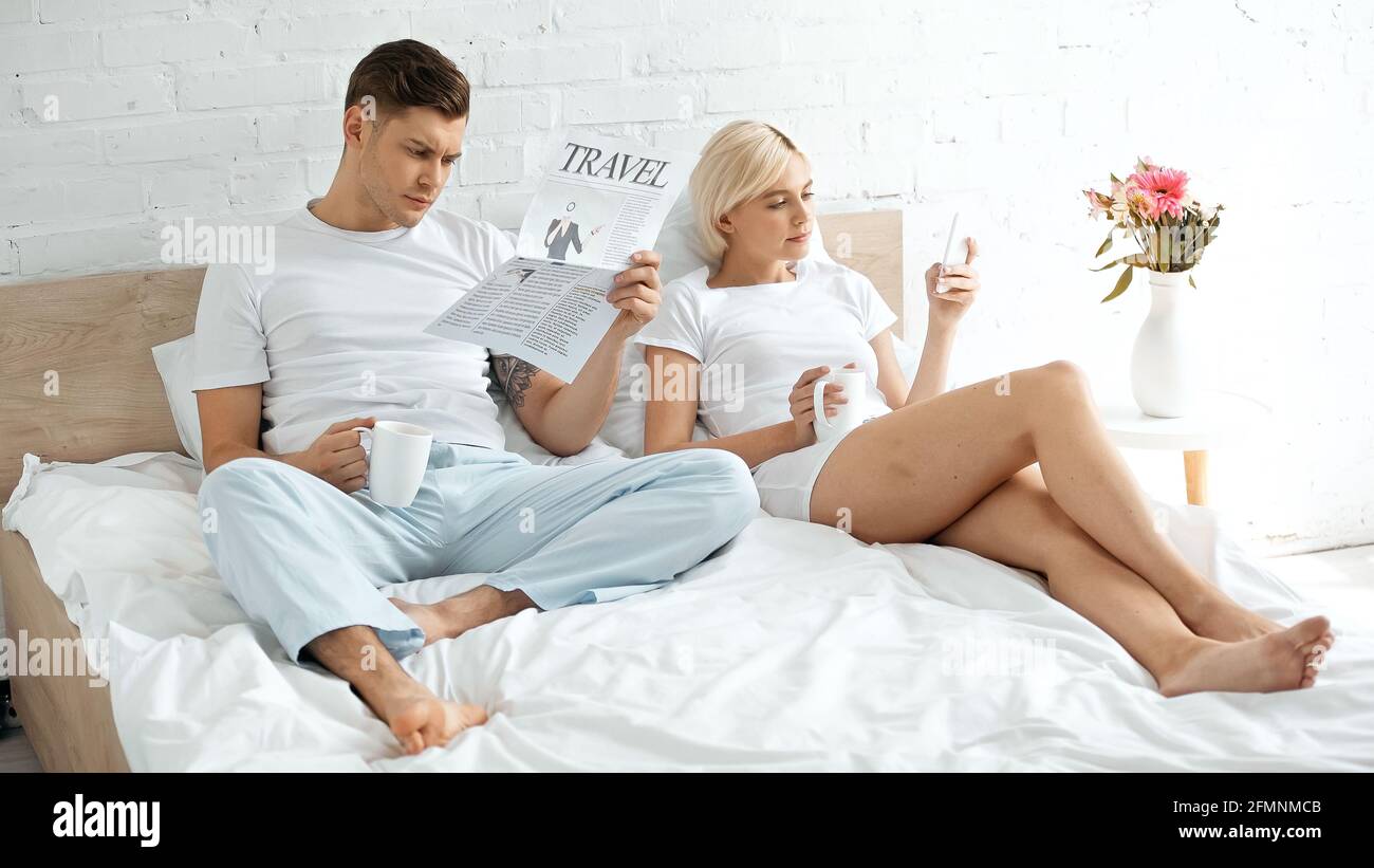 Tätowierte Mann Lesung Reise Zeitung in der Nähe Frau hält Tasse Kaffee und mit dem Smartphone im Bett Stockfoto