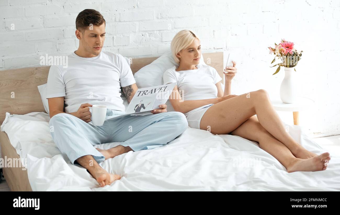Tätowierter Mann, der die Reisezeitung liest und eine Tasse Kaffee hält Frau in der Nähe, die Smartphone im Bett benutzt Stockfoto