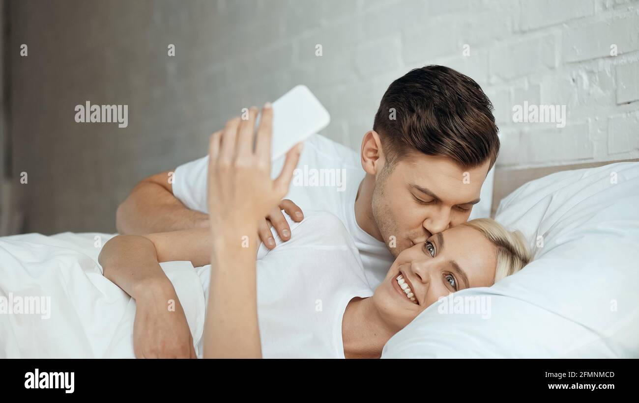 Junger Mann küsst Frau nimmt Selfie auf Smartphone im Bett Stockfoto