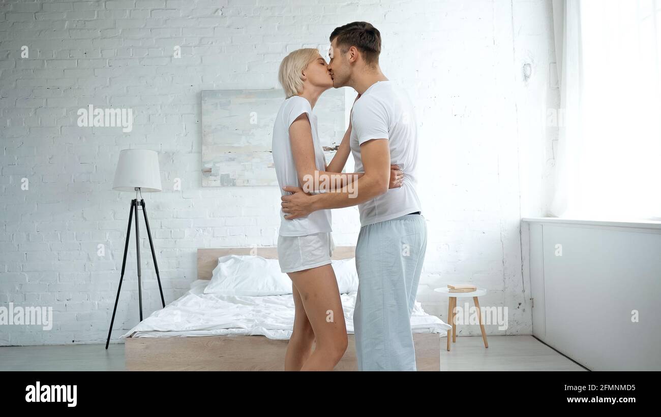 Seitenansicht des glücklichen Mannes, der die blonde Frau umarmt und küsst Im Schlafzimmer Stockfoto