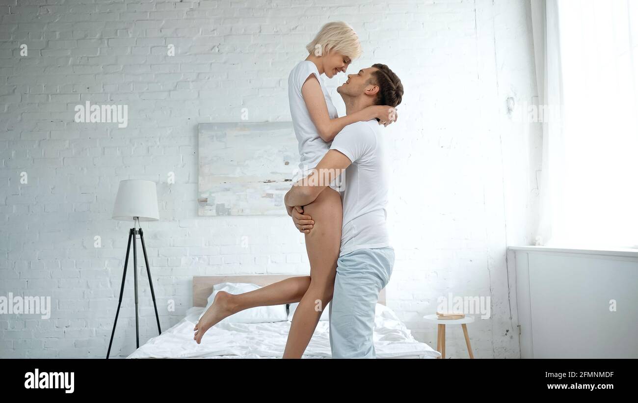 Seitenansicht des fröhlichen Mannes, der die blonde Frau in den Armen hält Im Schlafzimmer Stockfoto
