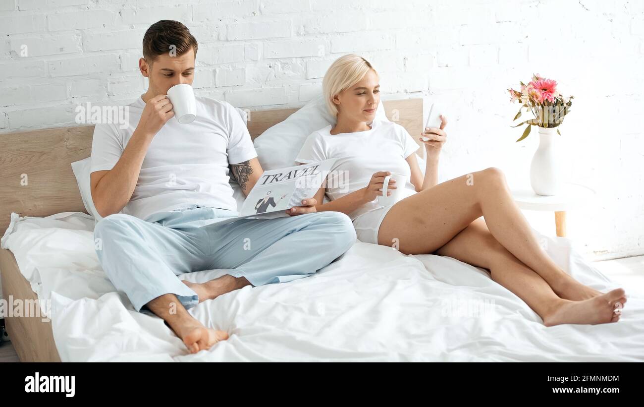 Tätowierter Mann, der in der Nähe einer Frau eine Reisezeitung liest und Kaffee trinkt Mit dem Smartphone im Bett Stockfoto