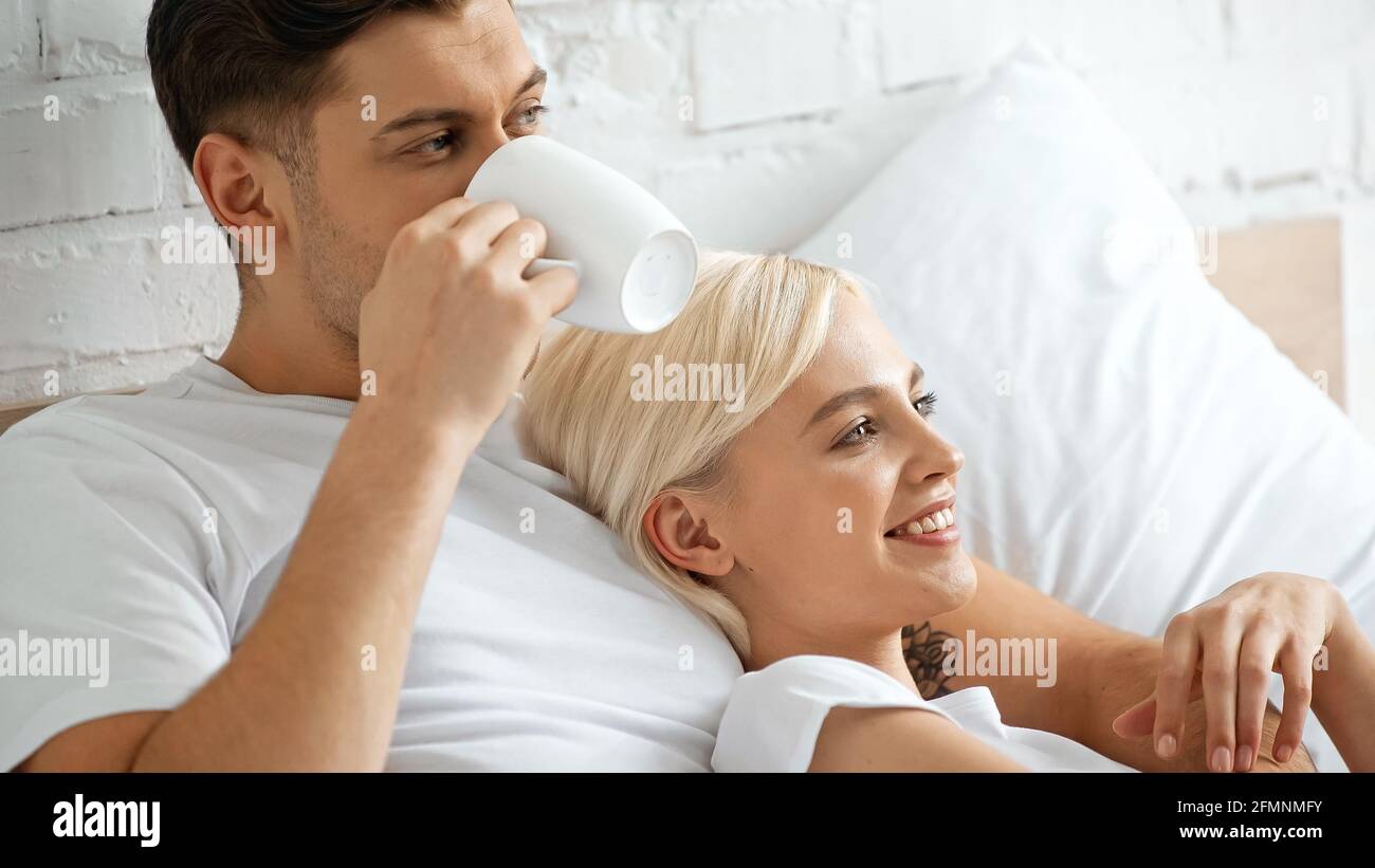 Tätowierte Mann, der Kaffee trinkt, während er mit einer angefreuten blonden Frau liegt Im Bett Stockfoto