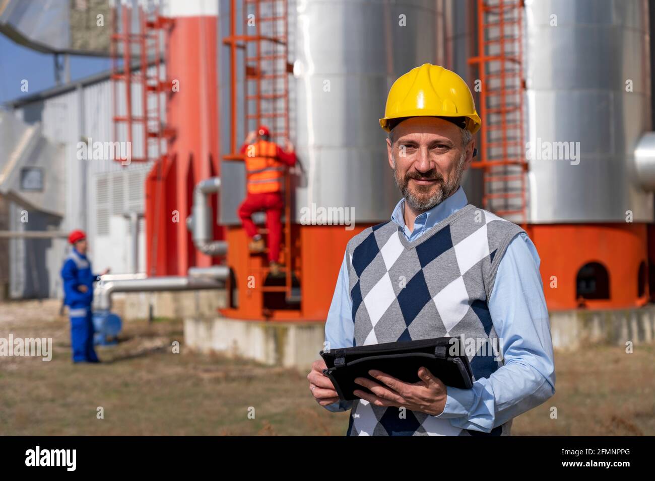 Ölingenieur mit digitalem Tablett, das vor den Lagertanks der Ölraffinerie steht. Porträt des Geschäftsmunters in gelbem Arbeitshelm. Stockfoto