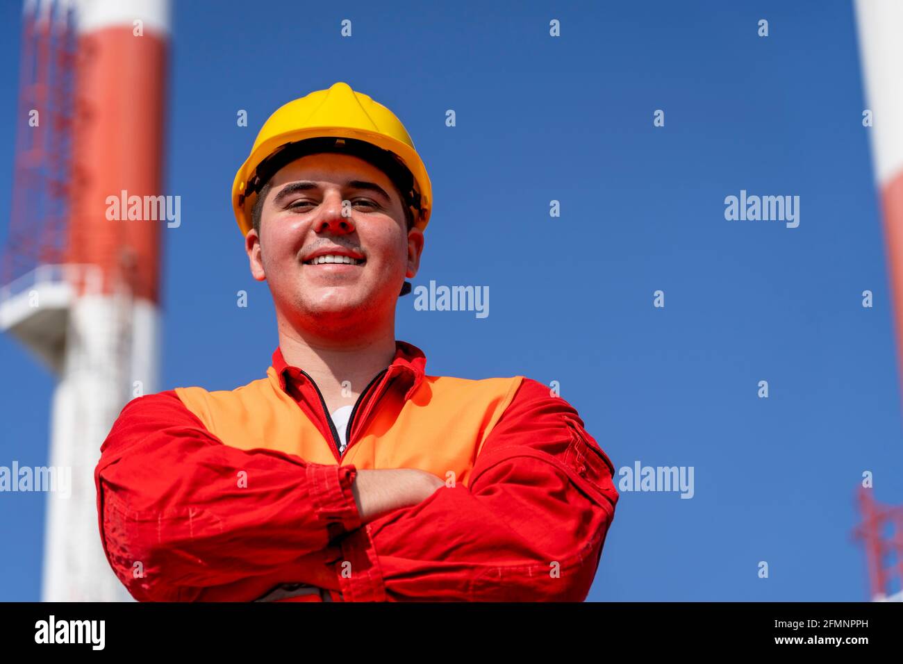 Porträt eines jungen Arbeiters in Red Coveralls und Yellow HardHat gegen die Schornsteine des Kraftwerks und den blauen Himmel. Lächelnder Arbeiter in schützender Arbeitskleidung. Stockfoto