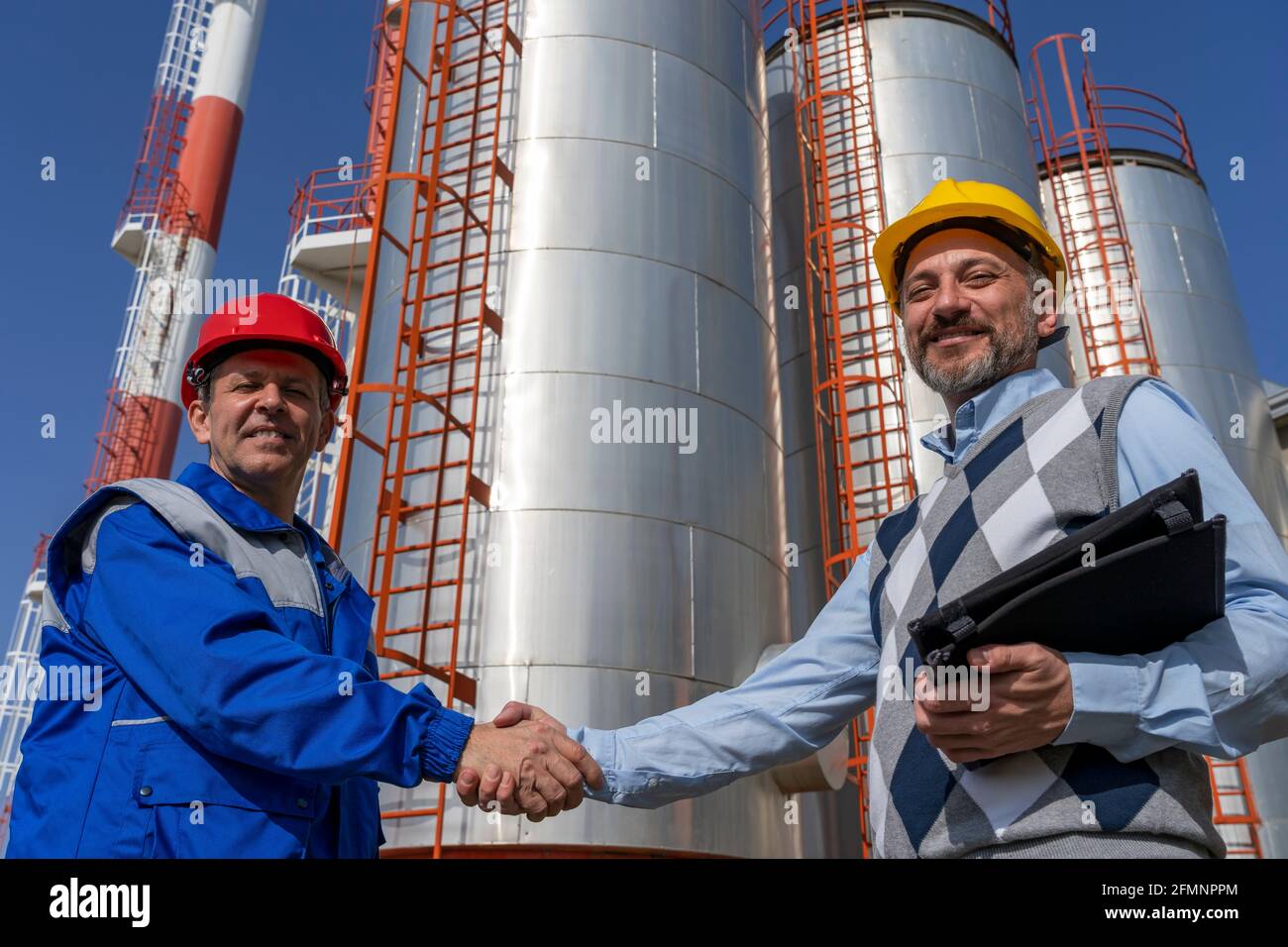 Lächelnder Arbeiter und Geschäftsmann schüttelten sich die Hände gegen Lagertanks für Ölraffinerien. Arbeiter in Arbeitsschutzkleidung Treffen mit Geschäftsleuten. Stockfoto