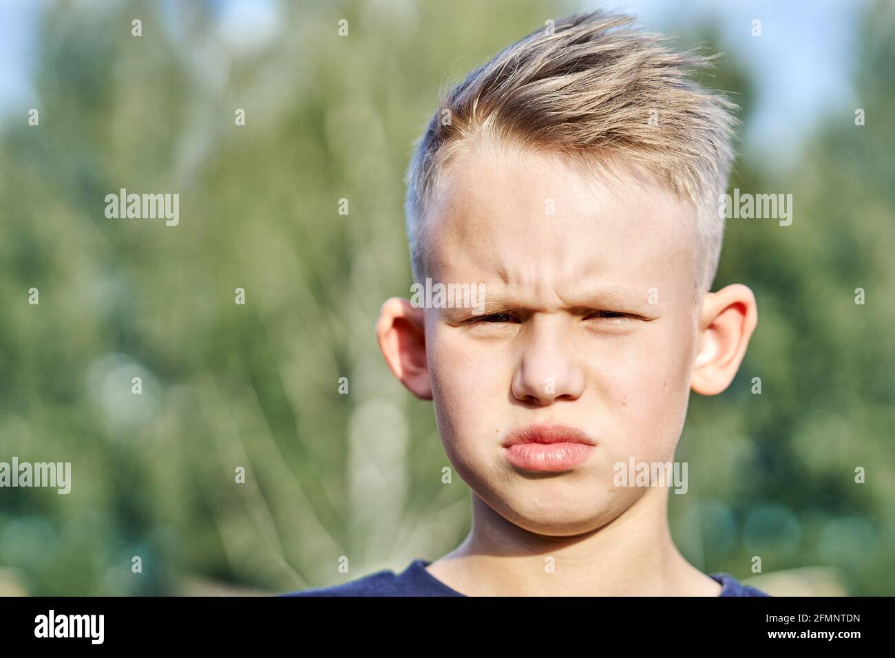 Frustrierter kleiner Schuljunge mit kurzer Frisur, der für die Kamera posiert Verschwommen grün sonnig Sommer Stadtpark extreme Nahaufnahme Stockfoto