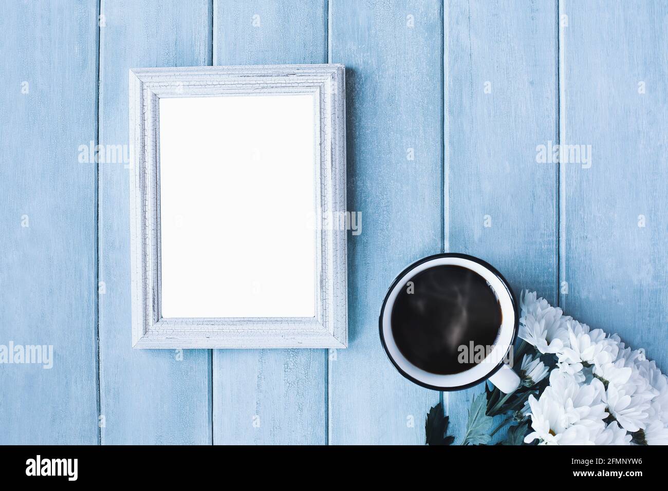 Leerer vertikaler Bilderrahmen auf blauem, rustikalem Hintergrund mit dampfendem Kaffee und weißen Gänseblümchen. Stockfoto