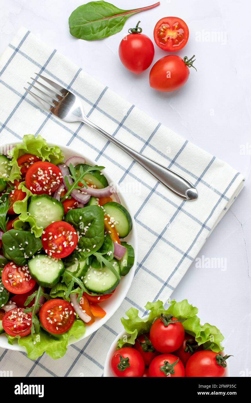 Gemüsesalat mit Kirschtomaten, Gurken, Rucola, Sesam, Basilikum und Olivenöl. Weiße karierte Serviette und Gabel auf einem Tisch. Ansicht f Stockfoto