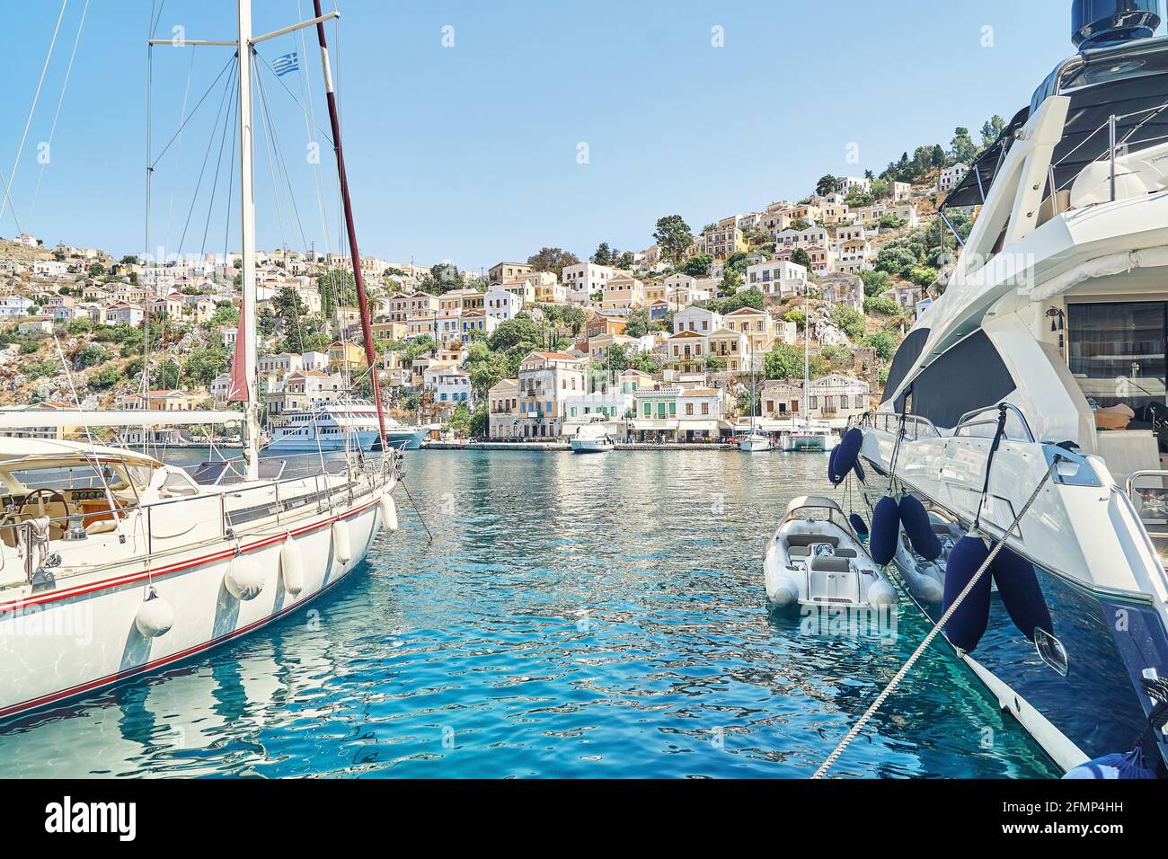 Festgemacht Segelboot und große Yacht auf dem Meer gegen historische Stadt Mit alten bunten Gebäuden, die auf Hügeln auf der Insel Symi verstreut sind In Griechenland Stockfoto