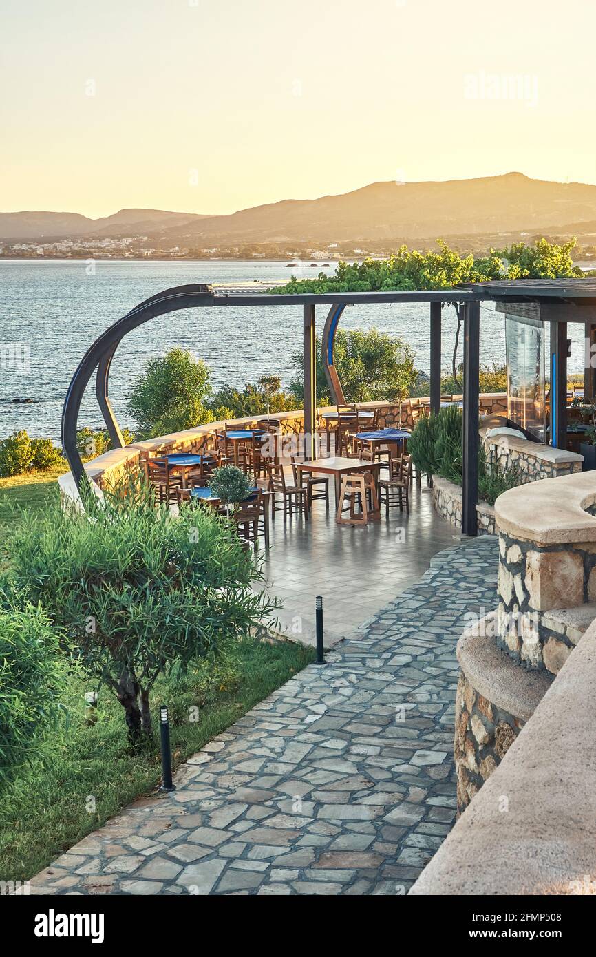Lokale Restaurant Terrasse mit Esstischen Stein gepflasterten Weg und Grüne Bäume vor blauem plätscherndem Meer bei Sonnenuntergang auf Rhodos Griechenland Stockfoto