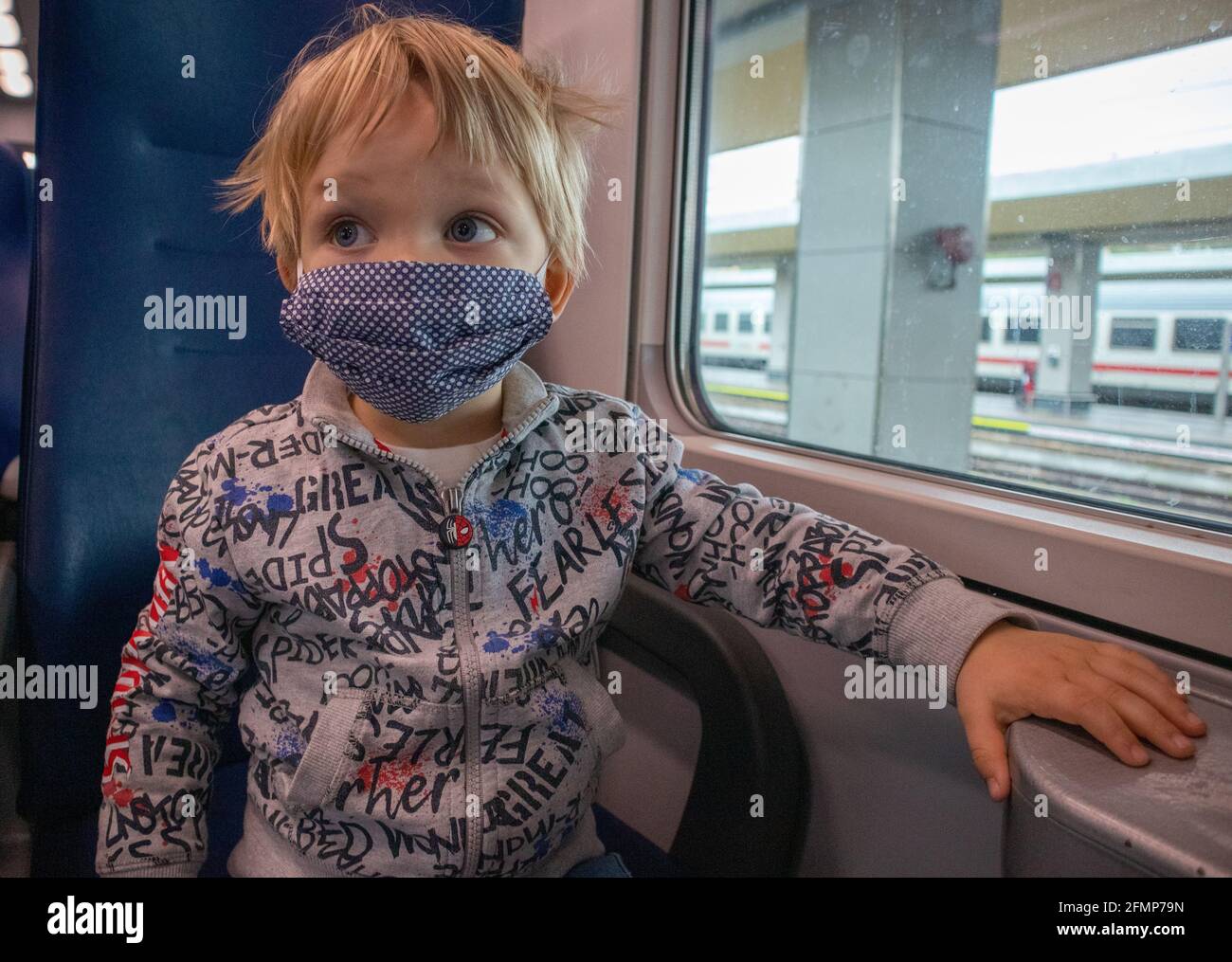 Blondes Kind mit blauen Augen und Gesichtsmaske, in der Nähe eines gelben Rucksacks, an Bord eines Regionalzuges in Italien Stockfoto