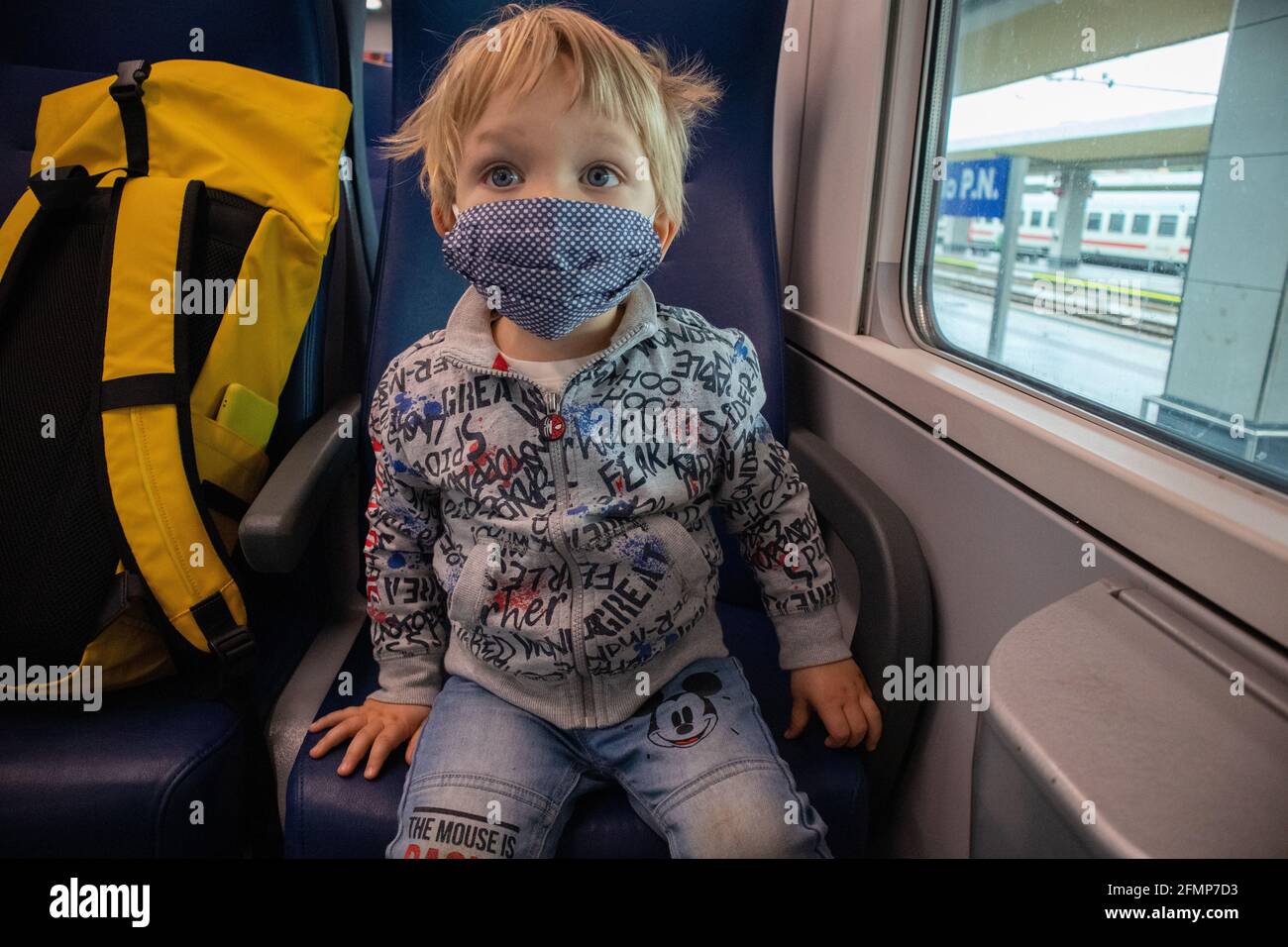 Blondes Kind mit blauen Augen und Gesichtsmaske, in der Nähe eines gelben Rucksacks, an Bord eines Regionalzuges in Italien Stockfoto