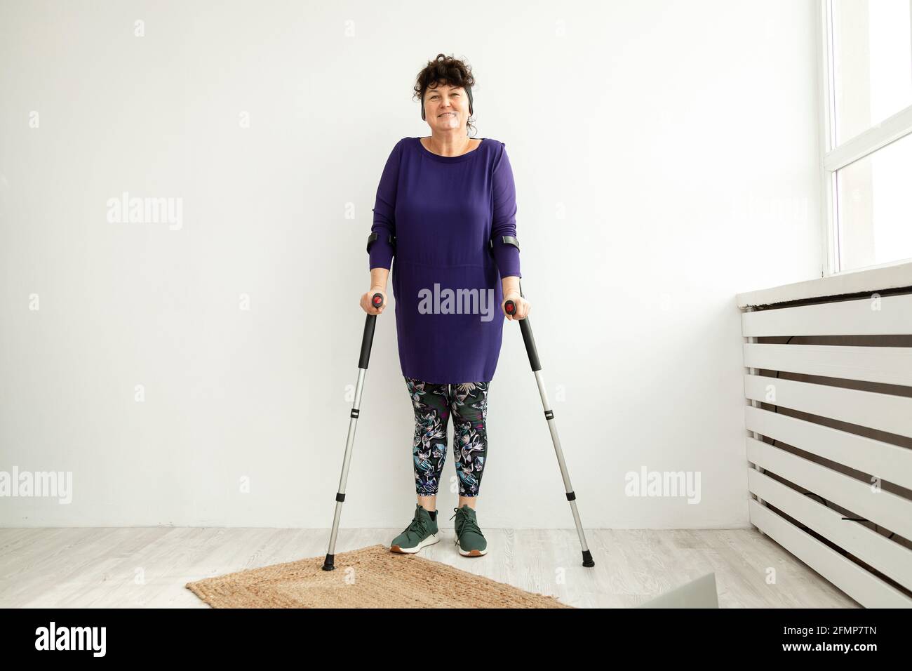 Frau im Alter steht auf weißem Hintergrund auf Krücken im Büro der Physiotherapeutin. Traumaerholungs- und Rehabilitationskonzept Stockfoto