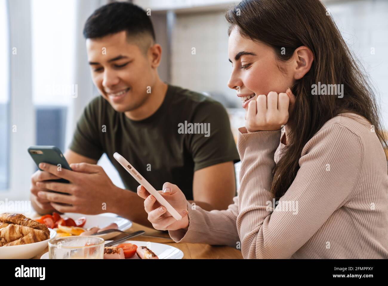 Portrait eines lächelnden Paares mit Smartphones und Gespräch beim Frühstück in der gemütlichen Küche zu Hause Stockfoto