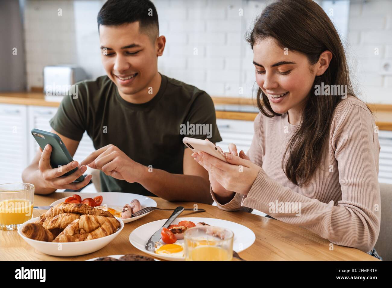 Portrait eines lächelnden Paares mit Smartphones und Gespräch beim Frühstück in der gemütlichen Küche zu Hause Stockfoto