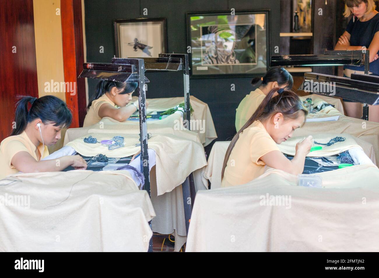 Junge vietnamesische Mädchen nähen eifrig Handstickerei auf Stoff, während die westliche Chefin in in der Ecke Hoi an, Vietnam, streng über sie wacht Stockfoto